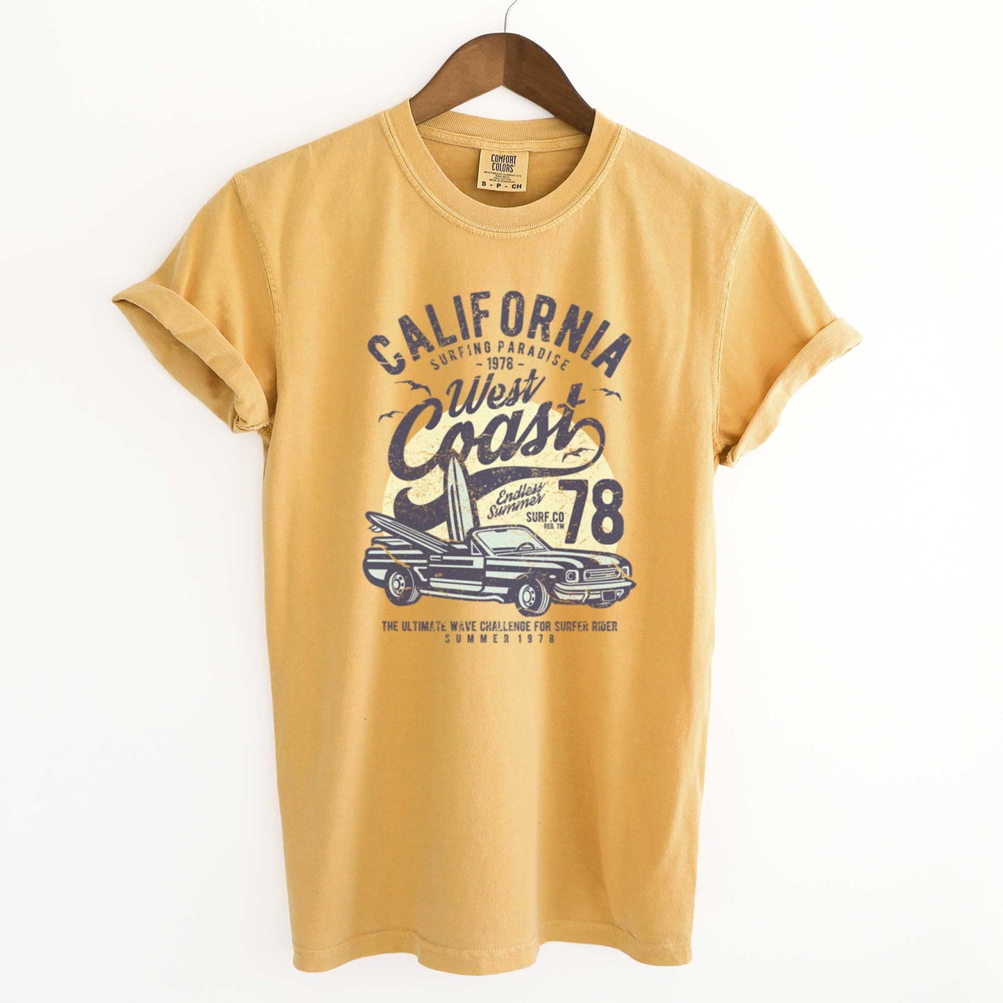 A hanging mustard yellow Comfort Colors t-shirt with surfboards inside a convertible and the words California west coast surfing paradise