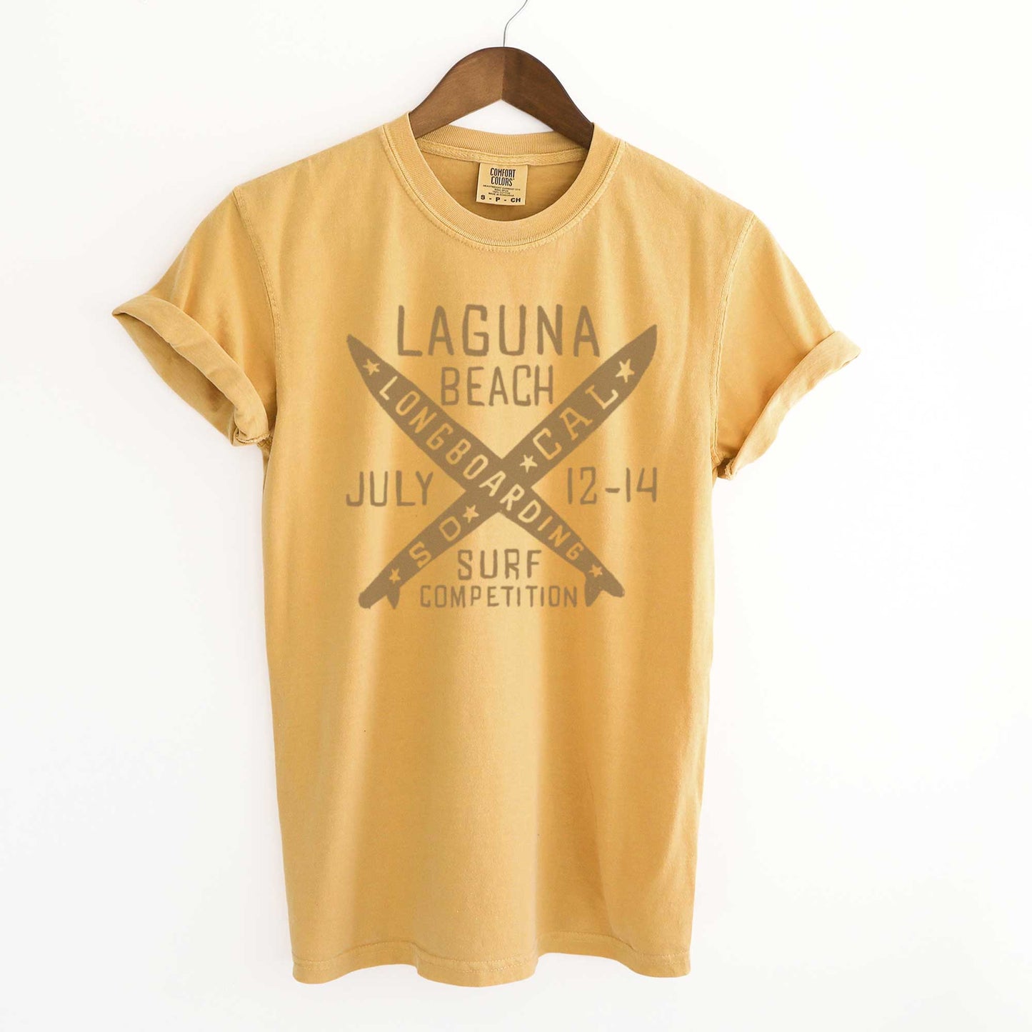 A hanging mustard yellow Comfort Colors t-shirt featuring 2 crossed longboards and the words Laguna Beach surf competition in brown ink