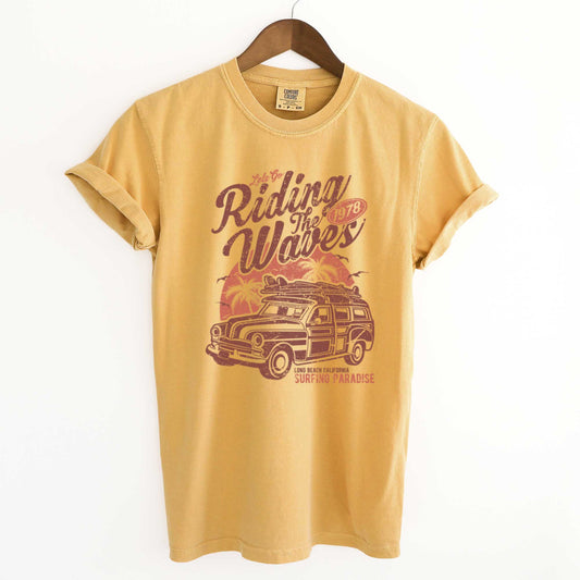 A hanging mustard yellow Comfort Colors t-shirt featuring a woodie station wagon, a surfboard, palm tress and the words let's go riding the waves