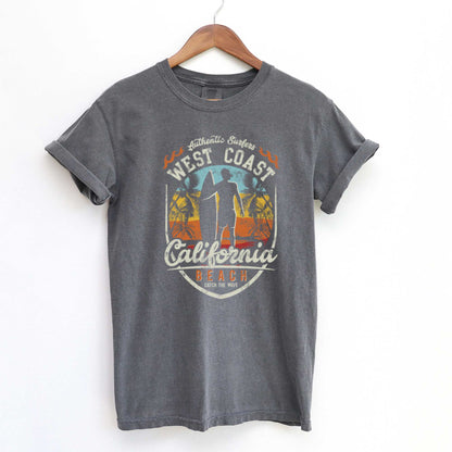 A hanging pepper gray Comfort Colors t-shirt featuring a man standing on a beach with a surfboard in front of a multi-colored sunset with the words California Beach.