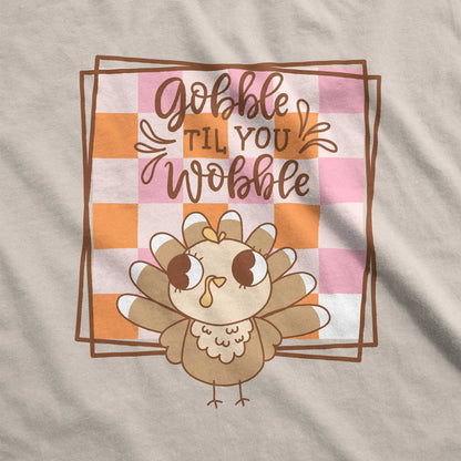 A  sand Gildan swatch featuring a cute cartoon turkey and a fall checkered background with the words gobble til you wobble.