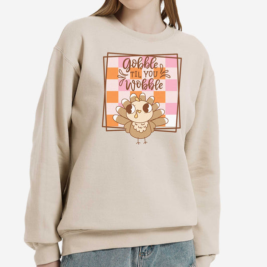A woman wearing a sand Gildan sweatshirt featuring a cute cartoon turkey and a fall checkered background with the words gobble til you wobble.