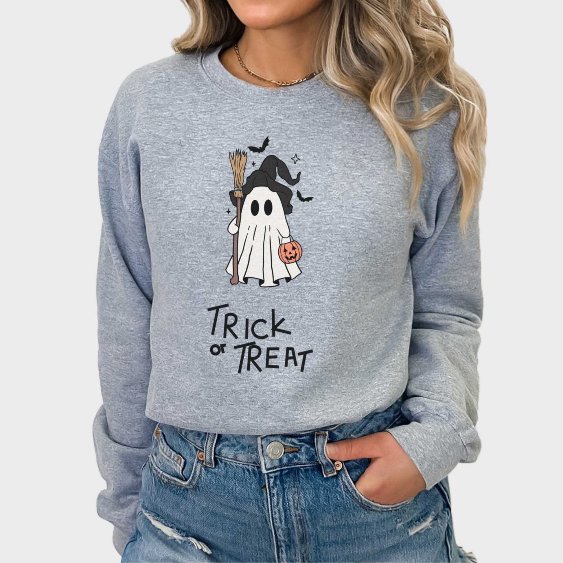 A woman wearing a sport grey Gildan sweatshirt featuring a cute cartoon witch ghost with the words trick or treat.