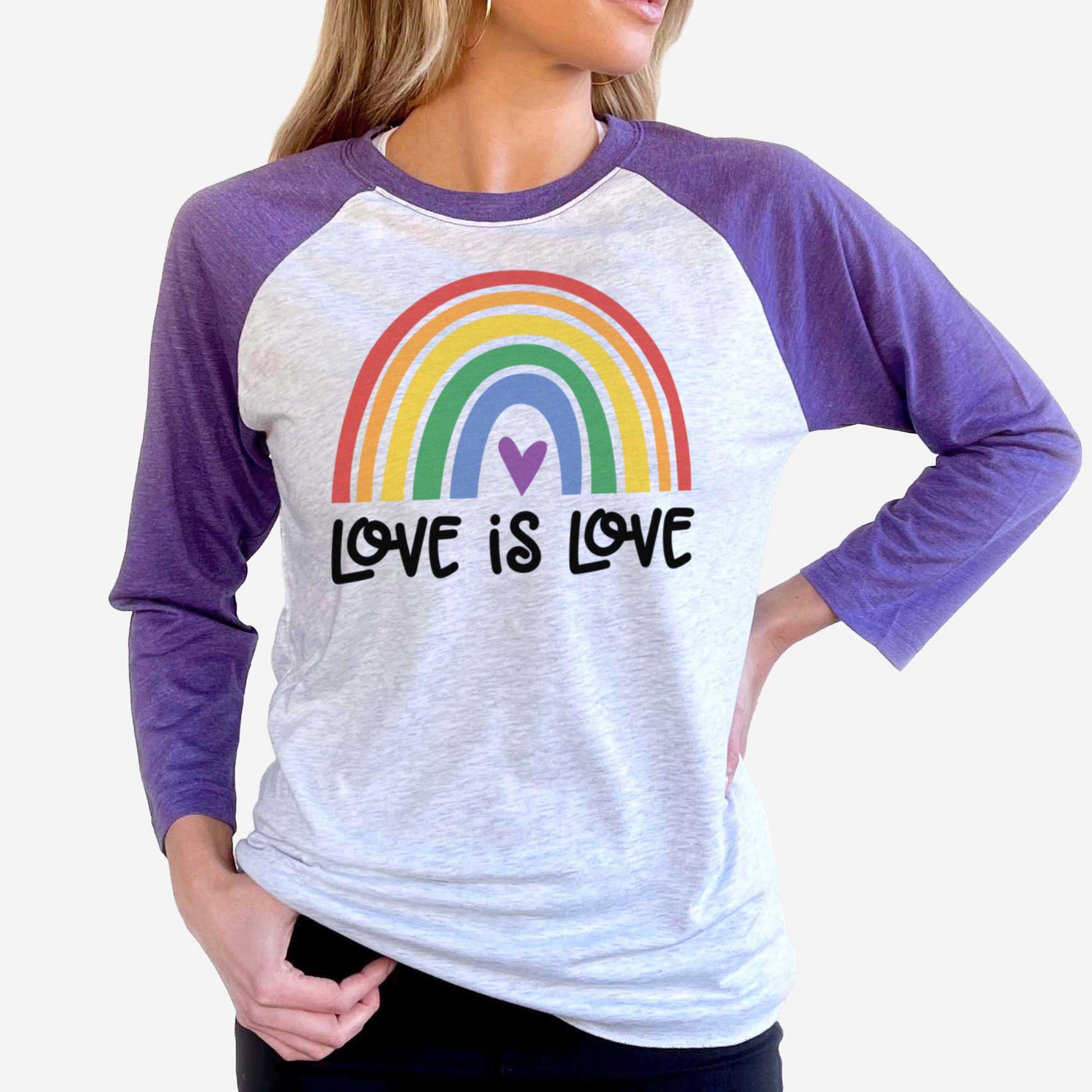 A woman wearing a purple rush and heather white Next Level raglan t-shirt featuring a multi-colored rainbow with the words love is love below it.