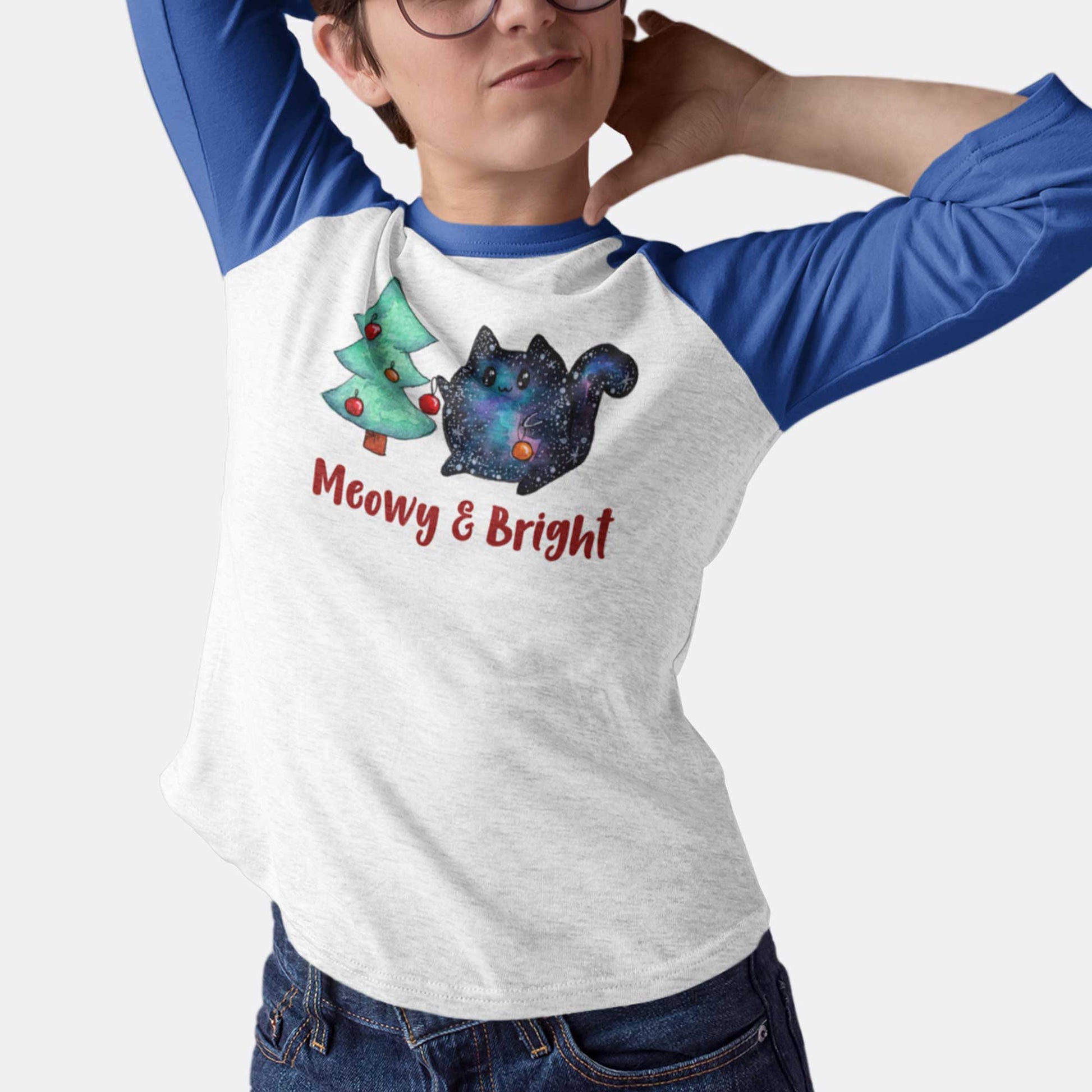 A woman wearing a vintage royal and heather white Next Level raglan t-shirt featuring a cartoon cat trimming a Christmas tree and the words meowy and bright.