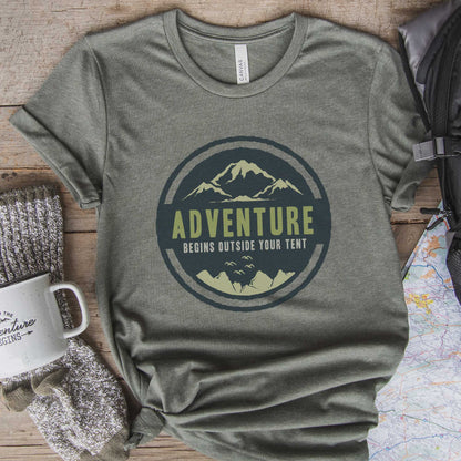 A heather military green Bella Canvas t-shirt surrounded by hiking gear that says adventure begins outside your tent.