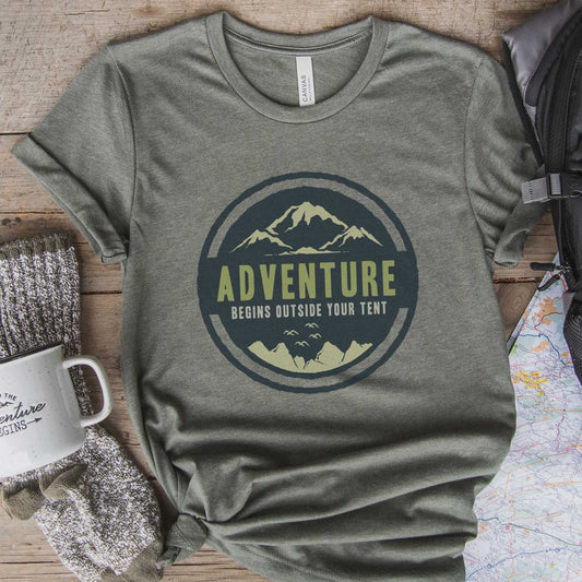 A heather military green Bella Canvas t-shirt surrounded by hiking gear that says adventure begins outside your tent.