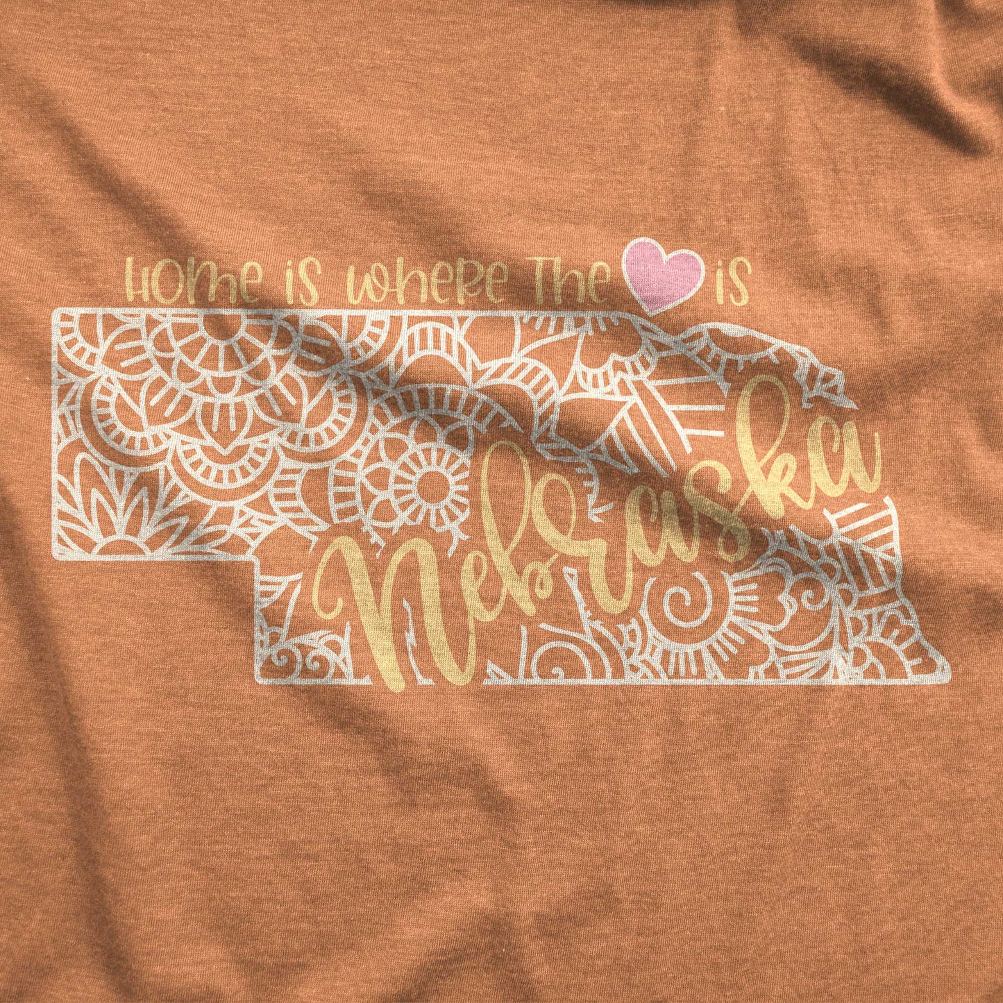 A heather autumn Bella Canvas swatch featuring a mandala in the shape of Nebraska with the words home is where the heart is.