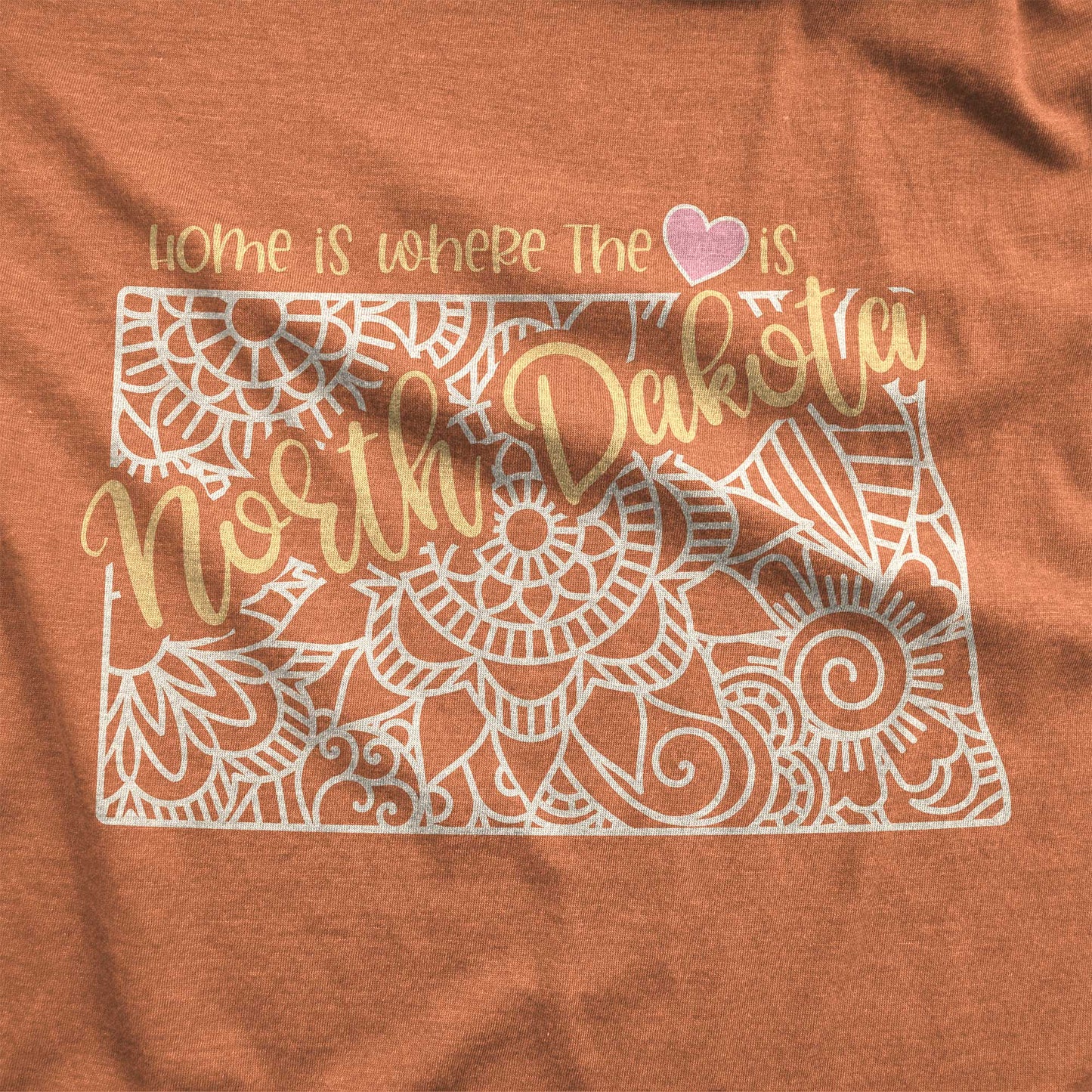 A heather autumn Bella Canvas swatch featuring a mandala in the shape of North Dakota with the words home is where the heart is.